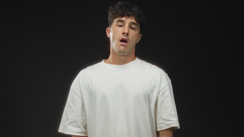 Man pointing finger to head in black studio wearing white t shirt, neutral pout and hand gesture toward temple, frontal portrait; sadness doubt.