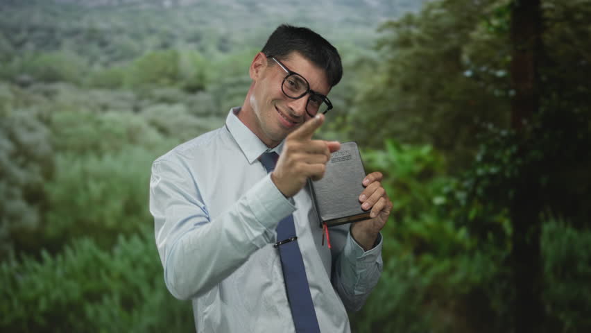 Man wearing light blue shirt and navy tie holds leather book to chest with warm smile in green park; faith serenity.