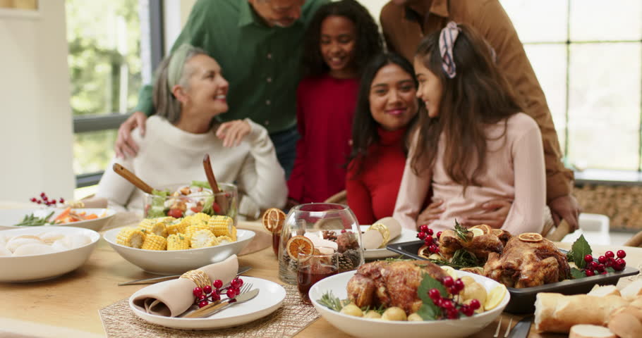 Diverse family leaning as grandmother gesturing to chicken, smiling and chatting at dining table. Family, celebration, togetherness, conviviality, rustic, warm, interior