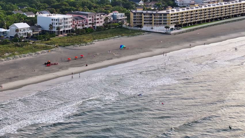 Panoramic view of Folly Beach near Charleston, South Carolina landscape showing beachfront homes and hotels during morning sunrise with people enjoying the shore and surfers in ocean water - 4K Drone