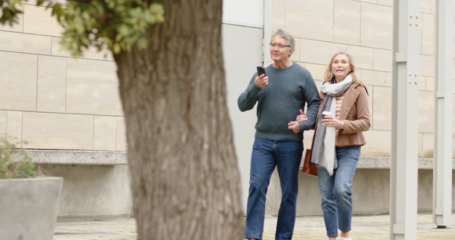 Senior couple emerging from left walking along walkway while holding smartphone and coffee, talking. Casual, conversation, outdoor, lifestyle, urban, connection, relaxation