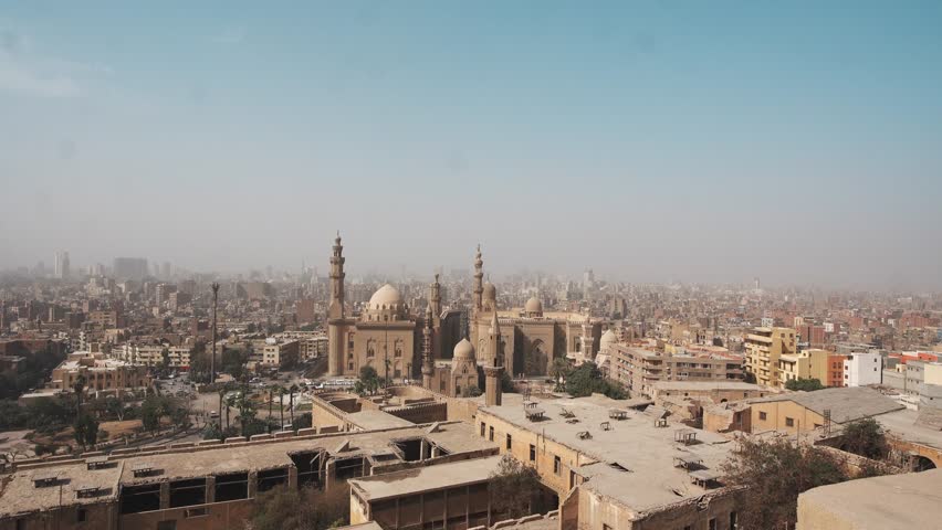 An aerial view of the urban cityscape in egypt features historic architecture and the iconic Sultan Hassan Mosque and Al Rifai mosque landmark under a clear blue cairo sky on a day