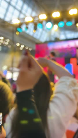 Crowd of fans cheering in a shopping mall atrium during a live concert. Young people filming the performance on smartphones in front of a bright stage. Energetic music event atmosphere with a singer performing for the audience.