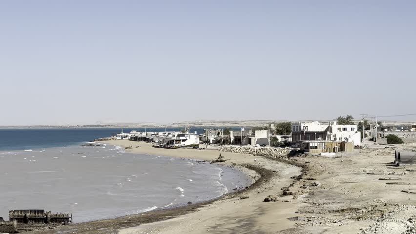 Coastal harbor shoreline with small boats and calm sea under clear blue sky, seaside waterfront landscape.
