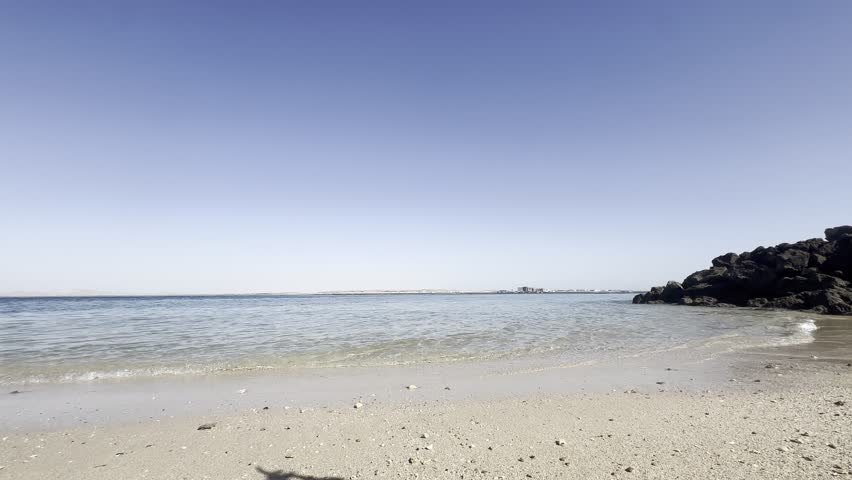 Calm sea and sandy beach with rocky shoreline
Wide shot of a calm blue sea meeting a sandy beach under a clear sky, with dark rocks on the shoreline. No people.
