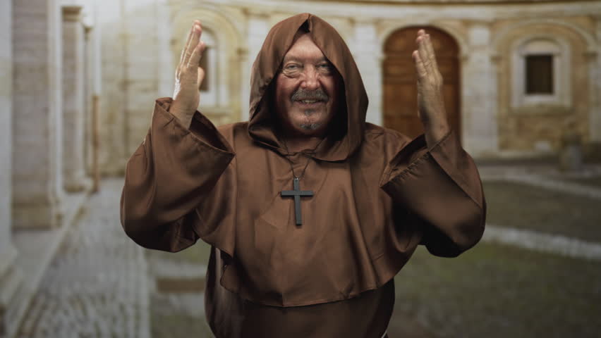 Man monk in brown habit with visible hands raised in prayer and cross necklace standing before a stone building courtyard; devotion prayer faith.