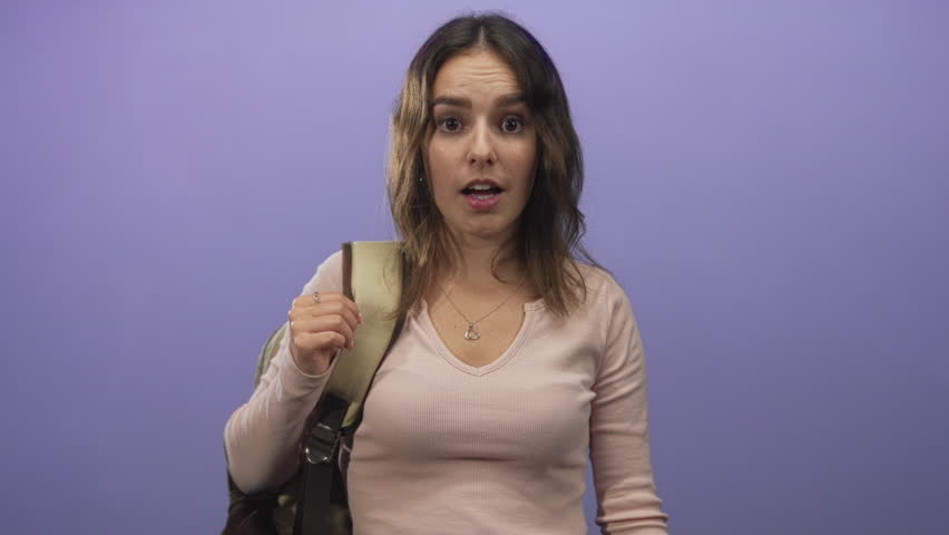 Young hispanic woman holding backpack strap with mouth open and heart necklace visible in studio purple backdrop; surprise new journey.