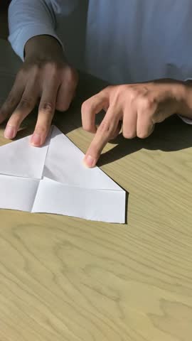 Close up in natural light of Asian man with cerebral palsy folding triangular paper in half and pressing firmly for paper airplane on wooden table. Fine motor skills training.