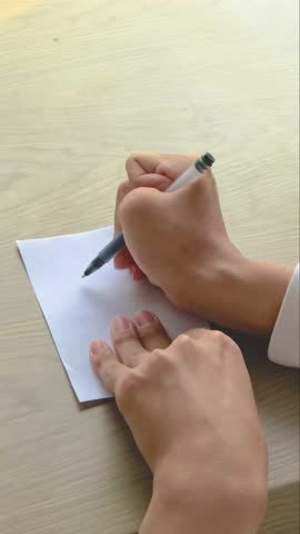 Close up in natural light of Asian man with cerebral palsy drawing triangle with right hand pen, left hand pressing paper on wooden table. Fine motor skills training.