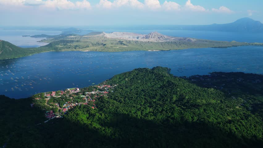 High-altitude drone shot showing Taal Volcano crater, surrounding mountains, Taal Lake, Agoncillo Baywalk, and geometric ocean fishpond grids with small boats.