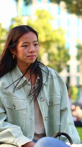 Candid portrait of a young asian woman with long dark hair, listening to someone off camera. She is sitting outdoors in a park on a sunny day