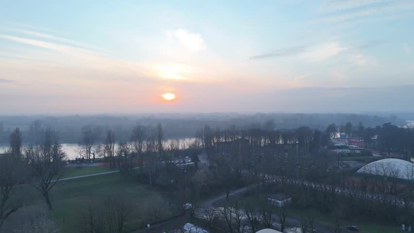 Sports fields and facilities near Po River in Cremona, Northern Italy, as the sun sets through light haze over bare trees, riverbanks, and the flat Po Valley landscape, wide aerial establishing shot