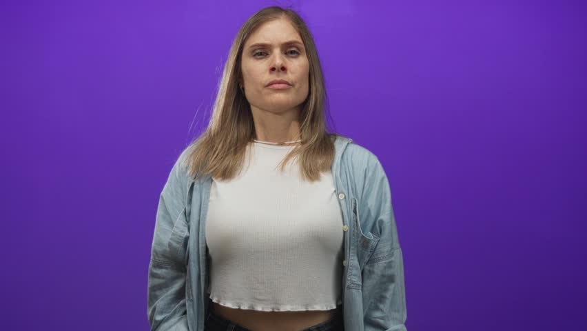 Woman clenches fists in purple studio wearing white crop top and denim shirt; determination strength.