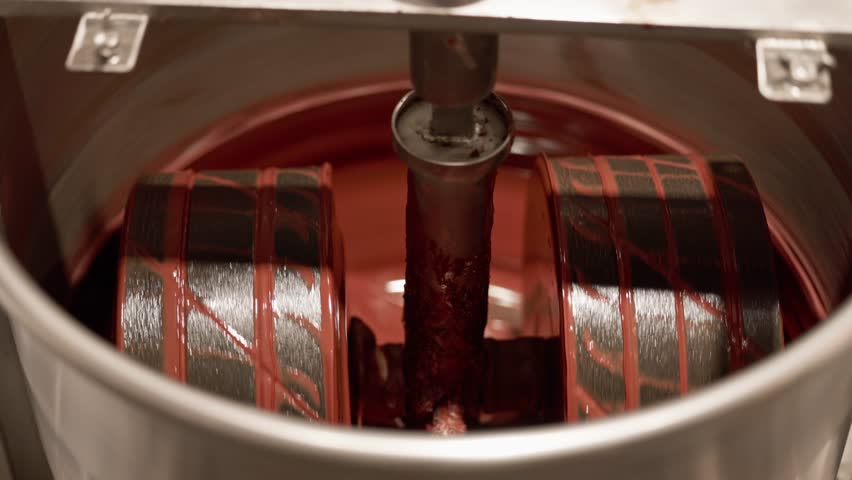Close up rotating industrial cocoa beans mixing rollers stirring and coating with glossy melted chocolate inside factory for confectionery production.