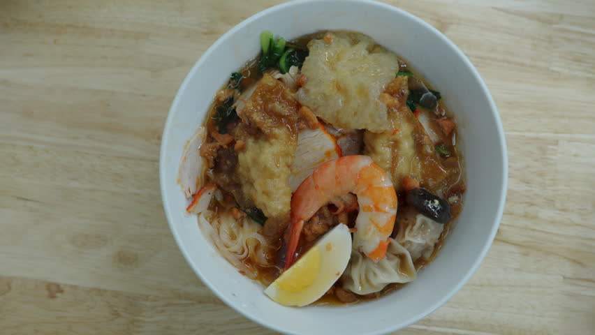 Wonton noodle soup bowl with shrimp and dumplings, Asian street food dish served with boiled egg and crispy topping, close up top view of traditional Chinese style noodle meal in ceramic bowl, savory broth with fresh ingredients on wooden table, authentic comfort food scene in casual restaurant setting, delicious handmade dumplings in hot soup