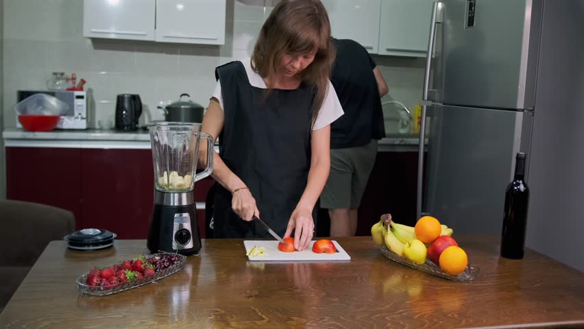 A woman in a black apron slices apples for a refreshing smoothie, while healthy ingredients such as strawberries, oranges and other fruits lie on the table and a man in the background makes breakfast.