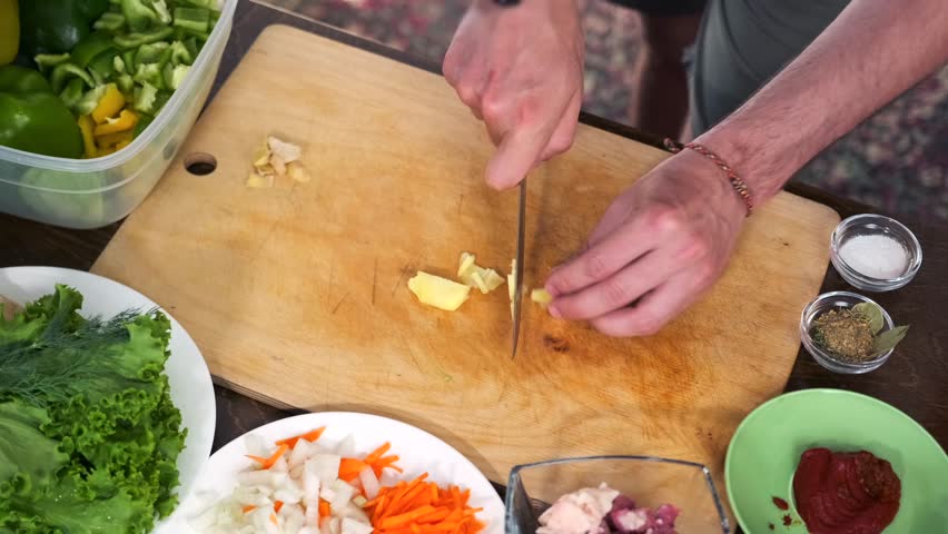 Male hands meticulously slice ginger on a cutting board, with plates of shredded carrots, onions, green and yellow peppers, lettuce leaves, a bowl of tomato sauce, and spices with pepper placed around