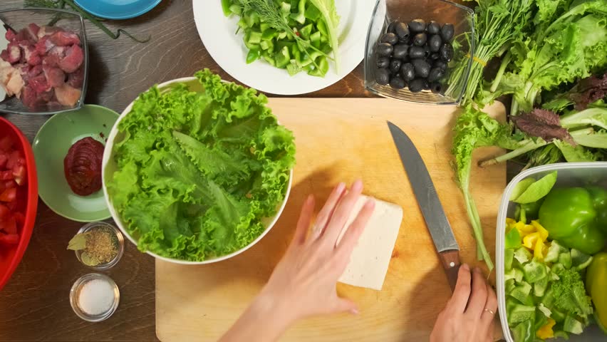 Top view of a woman hands cutting Greek feta cheese into cubes on a wooden board. Beside it is a bowl with crisp lettuce leaves, surrounded by black olives, fresh herbs, sliced vegetables, and spices.