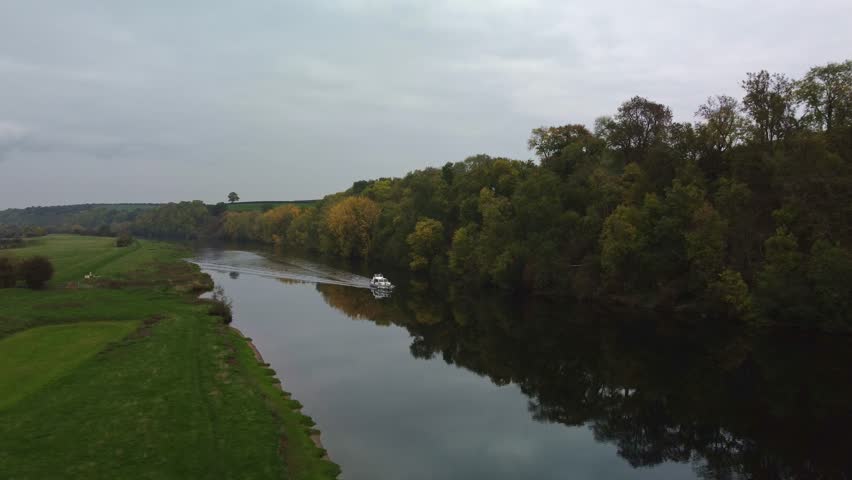 River boat travelling along the river Trent. Drone footage moves towards the boat and it gently travelling along the water causing peaceful waves along the fast water.