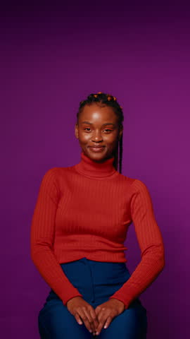 Young woman sits and smiles at the camera against a purple background in a vertical medium shot.