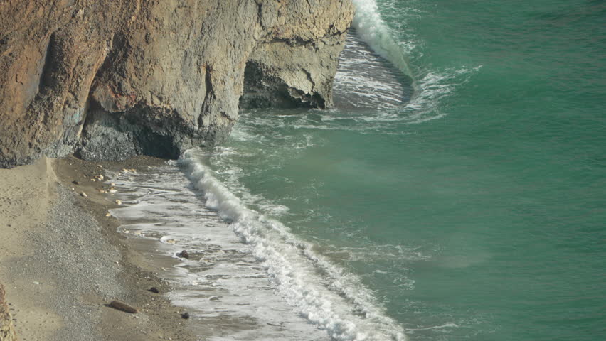 Ocean waves cliffside. Powerful turquoise sea water crashes onto a private pebble beach near a secluded rocky cave in slow motion.