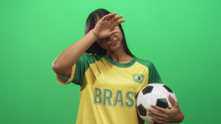 Young woman holds soccer ball, shields forehead and rests hand on hip in studio wearing brazil jersey; pride celebration.