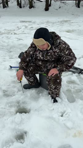 A man is sitting in the snow with a fishing pole and a bucket. He is wearing a hat and a jacket
