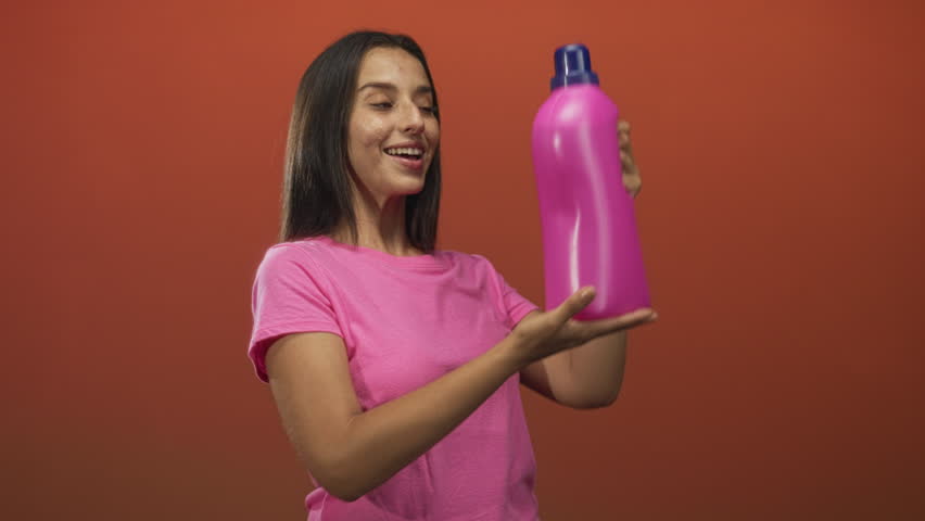 Woman holding large pink plastic bottle with handle while smiling in orange studio; joy product promotion.