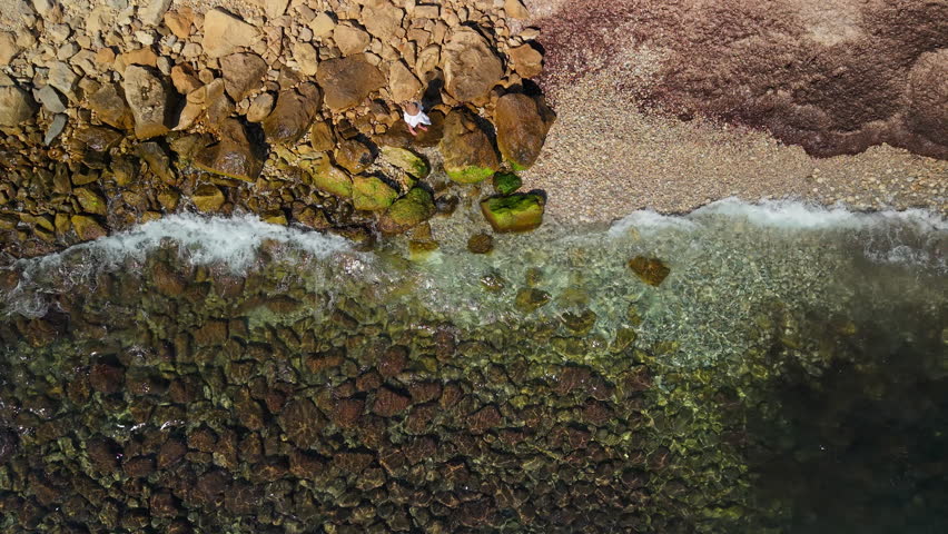 Aerial top view of middle aged woman standing on rocky Mediterranean shoreline with waves, travel freedom and solitude concept. High quality 4k footage