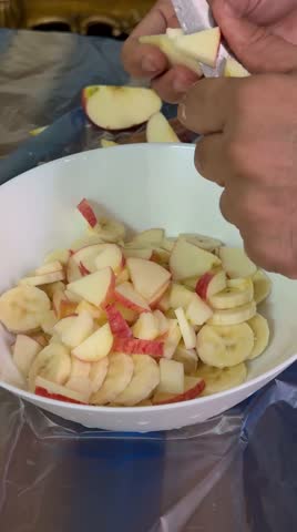 Close-up view of hands cutting fresh fruits and adding colorful fruit pieces into a white bowl to prepare healthy fruit chaat. Includes apple, banana, orange, and other fresh fruits. Bright clean background, healthy lifestyle concept, vegetarian food preparation, and fresh nutrition. Shot in 4K resolution.
