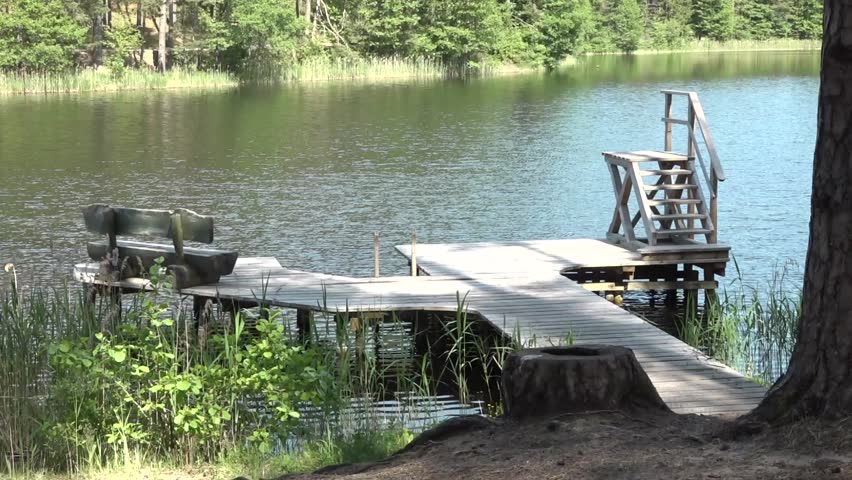 Wooden dock with a staircase and bench extends over a tranquil lake, surrounded by lush greenery, creating a peaceful outdoor atmosphere