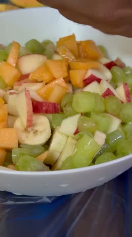 Close-up view of hands cutting fresh fruits and adding colorful fruit pieces into a white bowl to prepare healthy fruit chaat. Includes apple, banana, orange, and other fresh fruits. Bright clean background, healthy lifestyle concept, vegetarian food preparation, and fresh nutrition. Shot in 4K resolution.
