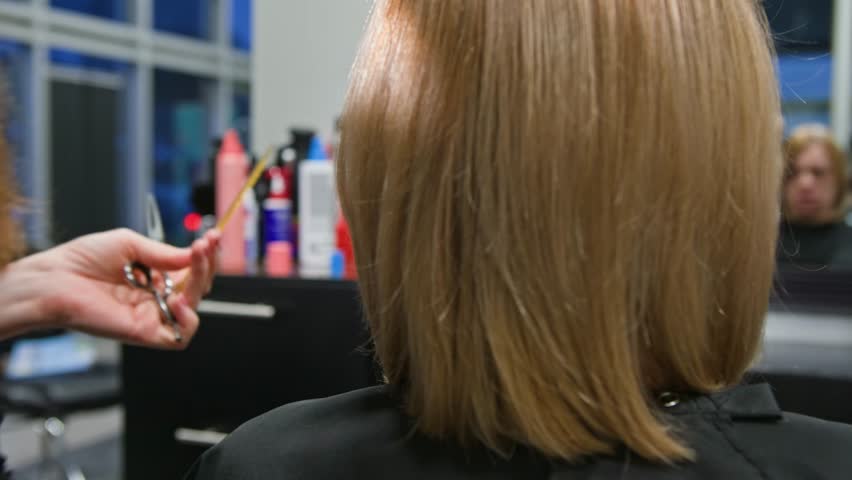 Woman sits in salon chair looking slightly worried as professional hairstylist cuts her hair length with scissors. Mirror reflection shows thoughtful expression during relaxing yet anxious haircut 