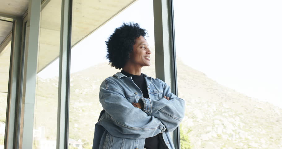 Spotting camera mid-adult man in jacket turning to it smiling laughing at home window facing hill. Contemplation, joy, relaxation, modern, architecture, casual, serenity