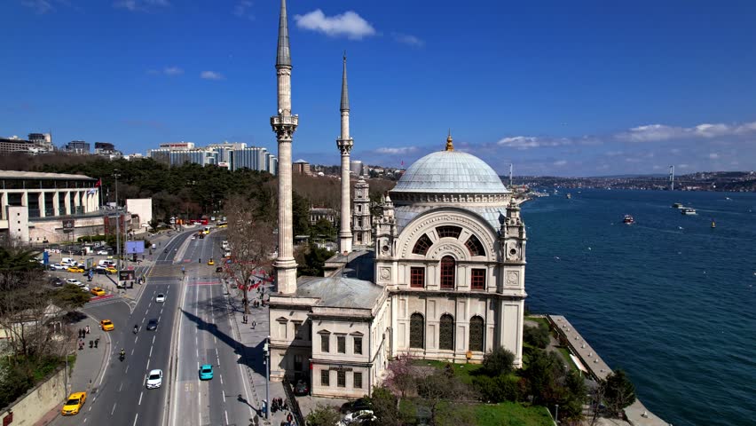 Wide-angle video frame of Dolmabahce Mosque by the Bosphorus in Istanbul, featuring its dome, minarets, sea traffic, coastal road, and the blend of historical and modern city life.