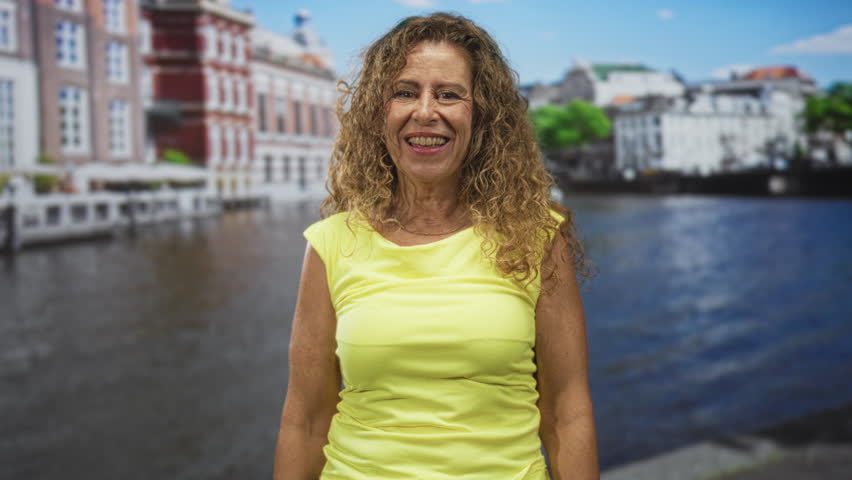 Woman covers mouth with hand by riverside building wearing yellow, surprised expression and open eyes; surprise playfulness warmth.