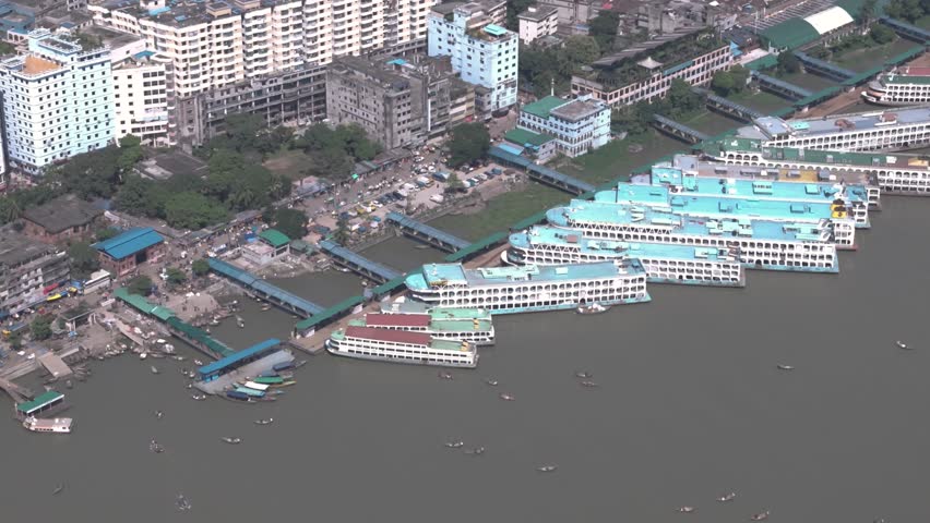 Cinematic aerial drone view of large passenger launch vessels docked at a busy river port terminal in Dhaka, Bangladesh. Urban waterfront with ferry transport, city buildings, and small boats on the river, showcasing transportation, infrastructure, and daily life in South Asia.