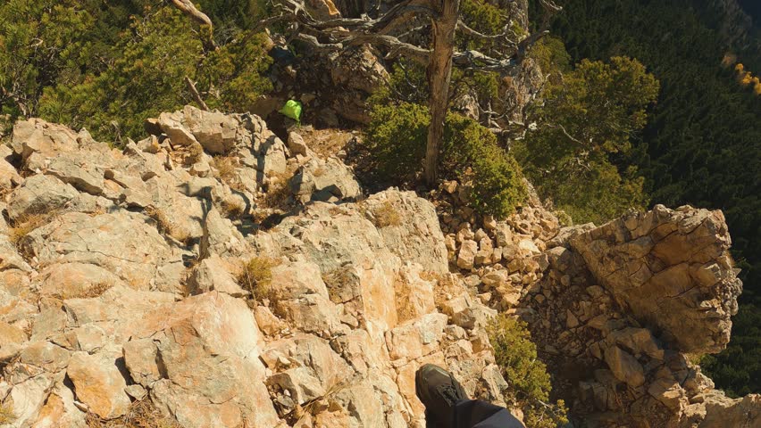A first-person perspective of a hiker descending rocks on a high, sunlit mountain. The dramatic, top-down angle creates a captivating sense of dizziness and adventure. Warm light