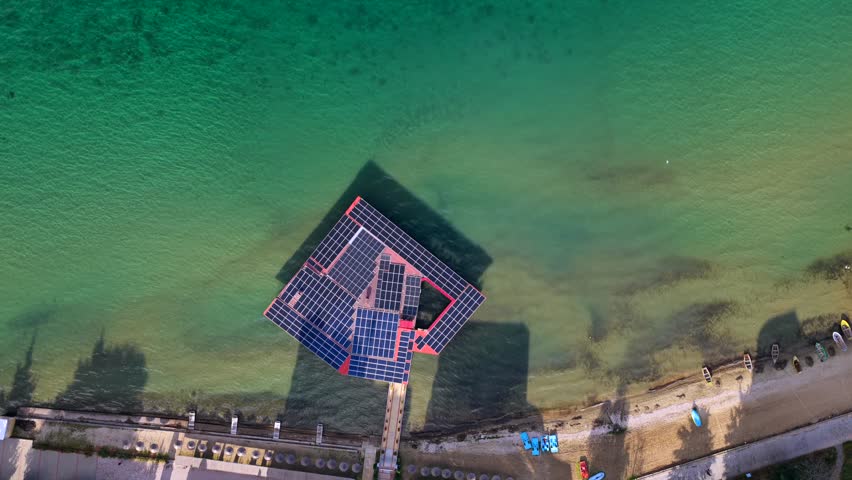 A top-down aerial video showing a modern solar-powered pier extending into the clear waters of Lake Ohrid in Pogradec. The video highlights geometric symmetry, solar panels, shallow shoreline textures, and small boats along the lakeside. Captured from a vertical drone perspective, this scene reflects sustainable design, clean energy, and the natural beauty of one of Albania’s most iconic freshwater lakes.