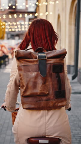 Back view of unknown young ginger woman in casual outfit, with backpack. Cycling by deserted city square decorated with lights, with open-air cafe. Autumn, urban lifestyle. Tracking shot, slow motion
