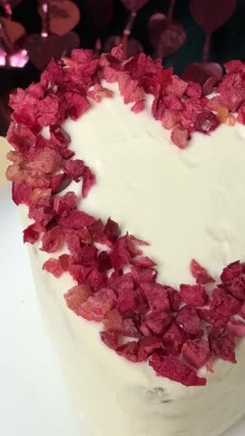 White heart cake with red decorations on top, standing on a white table against a blurred pink festive background. celebration, dessert, pink bokeh, Valentine’s Day
