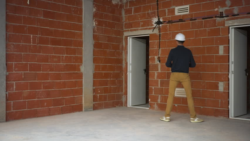 Male construction worker in hard hat approaches an open door while holding blueprints inside a partially finished building, featuring exposed brick walls and an unfinished floor throughout the space