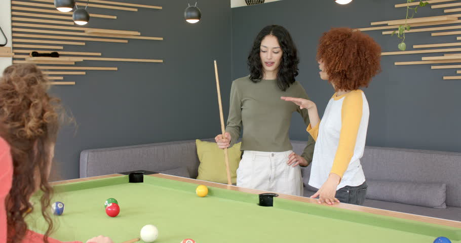 Female friends discussing angles at pool table, urging third striking cue pocketing ball, cheering. Recreation, socializing, game, entertainment, leisure, vibrant, teamwork