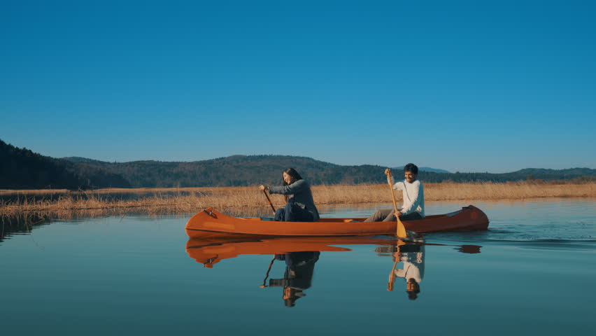 A couple paddles a canoe on a serene lake, surrounded by autumnal reeds and distant hills. The clear blue sky and calm water reflect their peaceful activity, capturing a moment of connection.