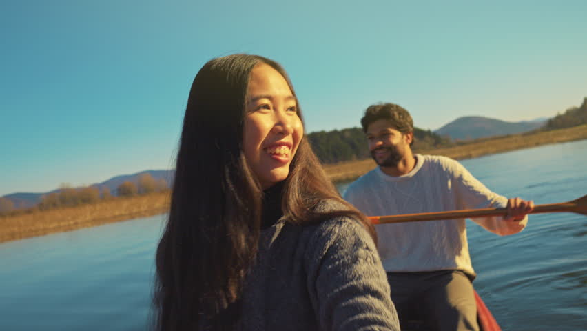 A young couple paddles a canoe on a serene lake, with the woman enthusiastically rowing and the man smiling behind her. The clear blue sky and distant mountains create a picturesque backdrop.