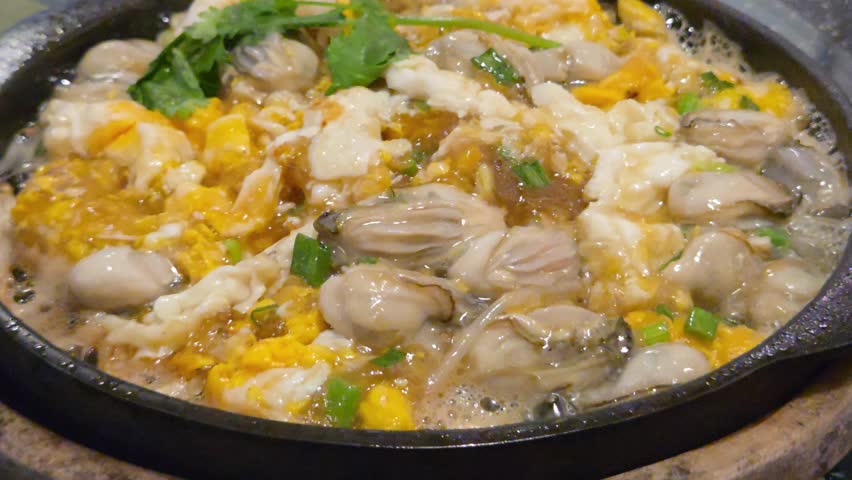 A Sizzling Hot Plate of Oyster Omelet (Hoi Tod) Featuring Fresh Oysters and Scrambled Eggs Garnished with Green Onions and Cilantro.	
