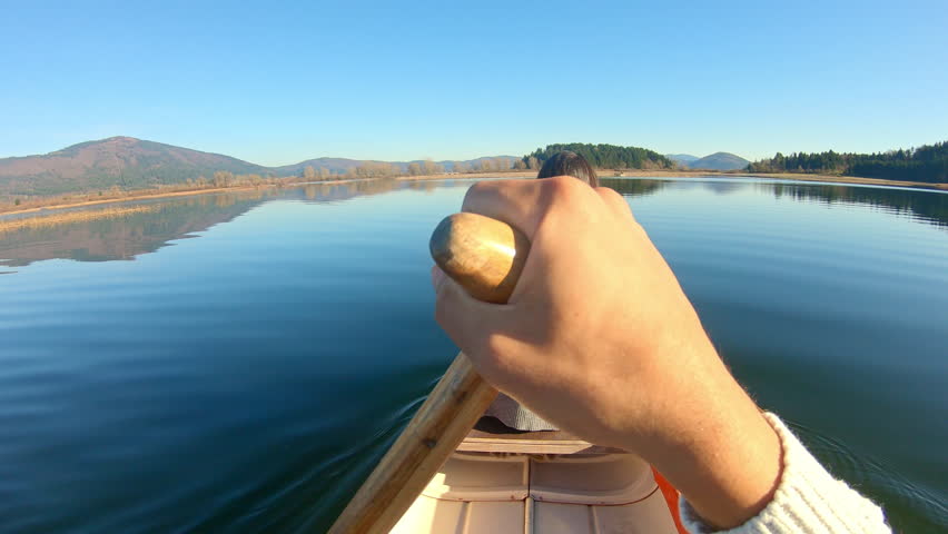 A woman paddles a canoe on a serene lake, surrounded by autumnal mountains. Her long hair flows as she raises her hand, capturing the moment.