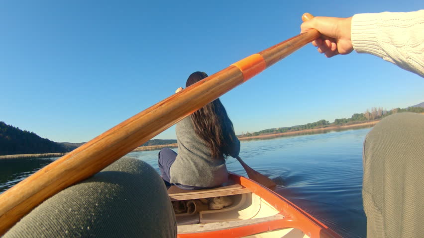 A couple paddles a canoe on a serene lake, surrounded by forested hills under a clear blue sky. The woman, with long dark hair, sits at the front, while the man, in a white sweater.