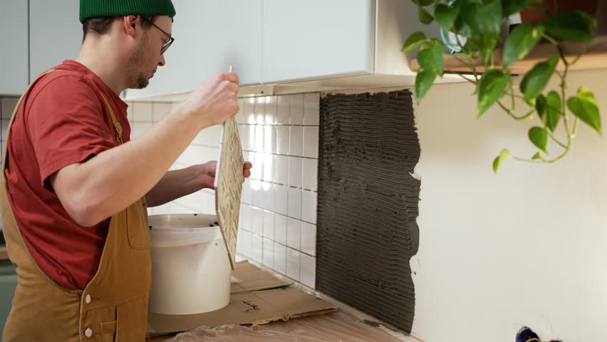 Professional tiler installing white ceramic tiles on a kitchen backsplash wall with adhesive. Home improvement and interior renovation process with a skilled construction worker