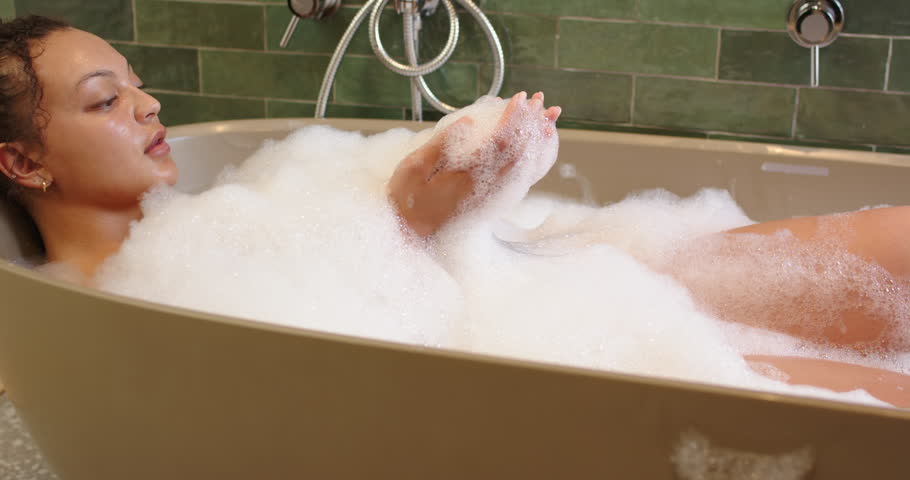 Foam on chest, African American woman reclining in tub, scooping and blowing bubbles, relaxing. Suds, bath, spa, relaxation, serenity, wellness, selfcare